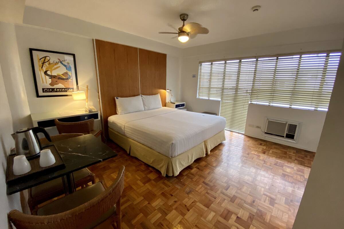 A bedroom with a bed, a desk, and a chair. There is a ceiling fan, a window with blinds, and a picture on the wall.