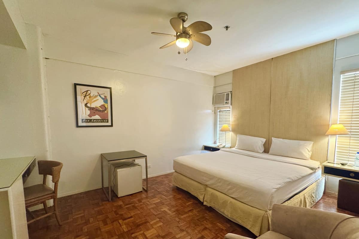A hotel room with a bed, a desk, a chair, a ceiling fan, and a framed picture on the wall.