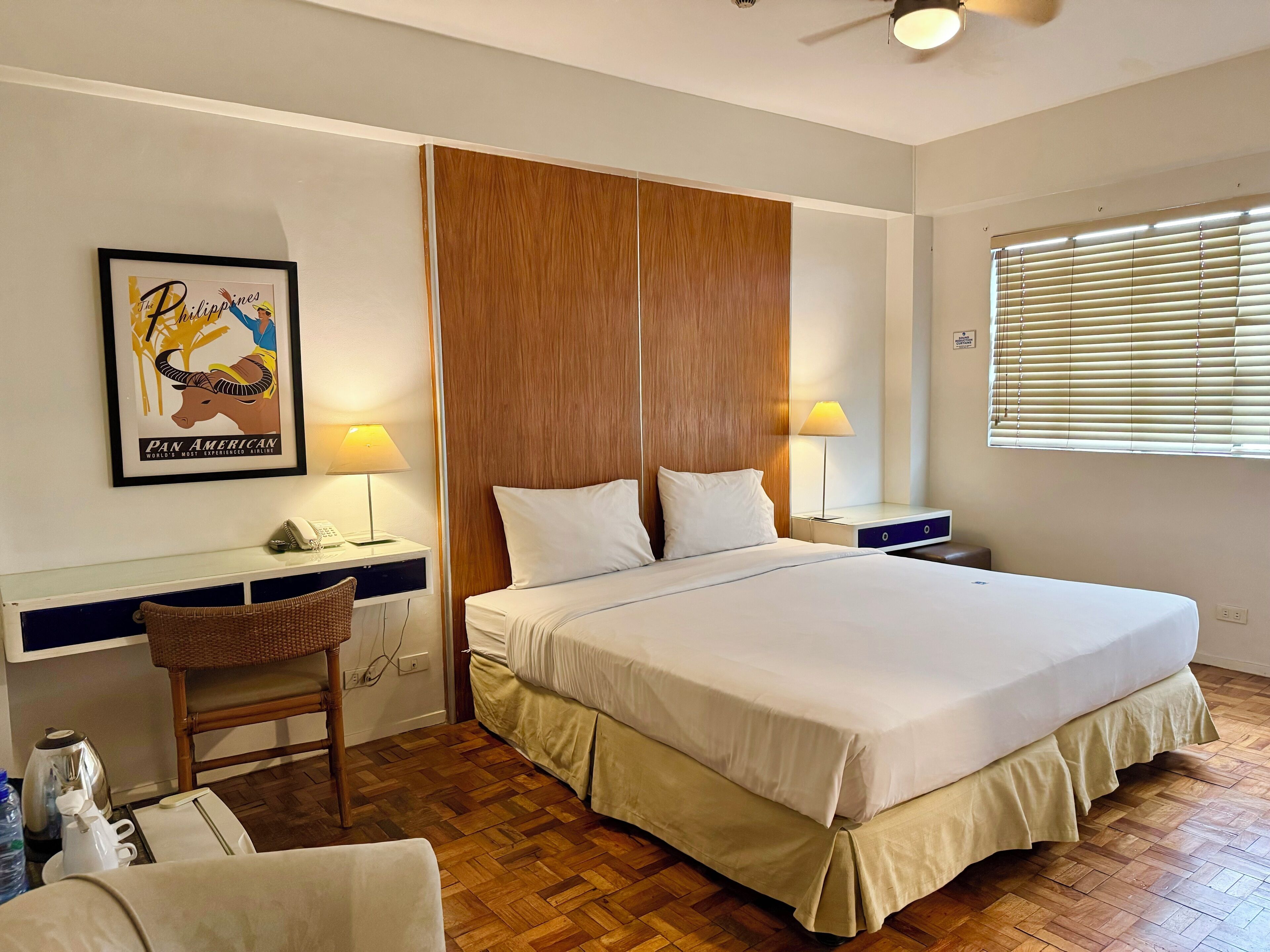A hotel room with a large bed, wooden headboard, two bedside lamps, a desk with a chair, a framed picture on the wall, and a window with blinds.