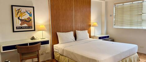 A hotel room with a large bed, wooden headboard, two bedside lamps, a desk with a chair, a framed picture on the wall, and a window with blinds.