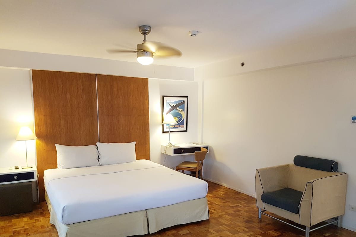 A hotel room with a large bed, a desk with a chair, a ceiling fan, and a wooden headboard.