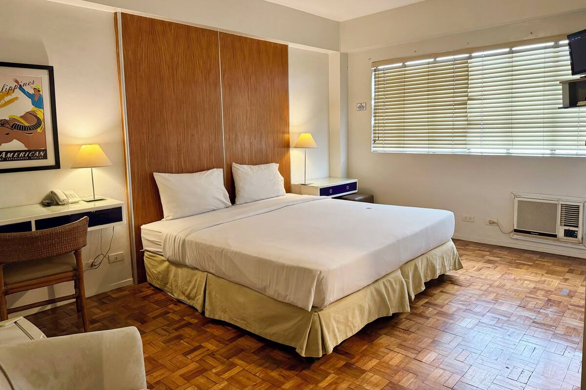 A hotel room with a large bed, wooden floor, two bedside lamps, a desk with a telephone, and a window with blinds.