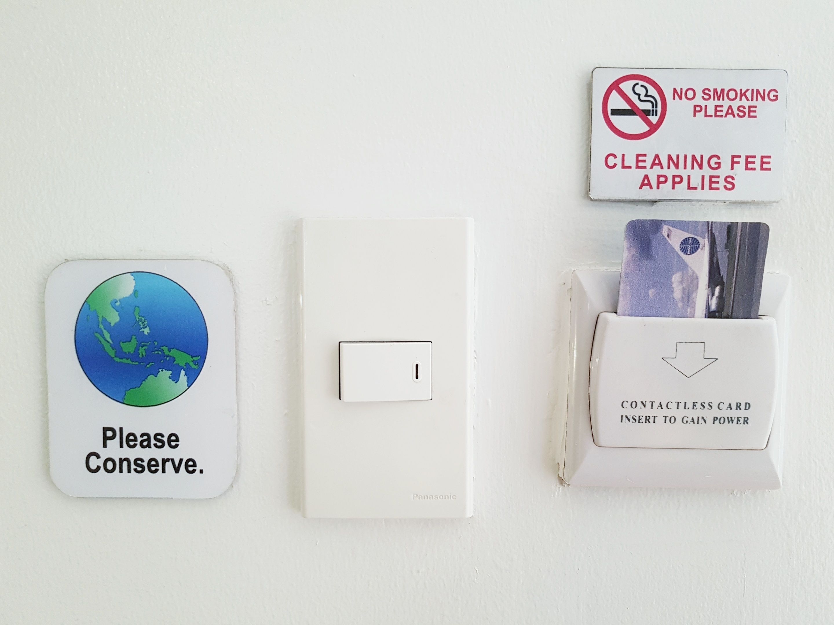 A wall with a globe sign saying "Please Conserve.", a light switch, a no smoking sign, and a card reader for contactless payment.