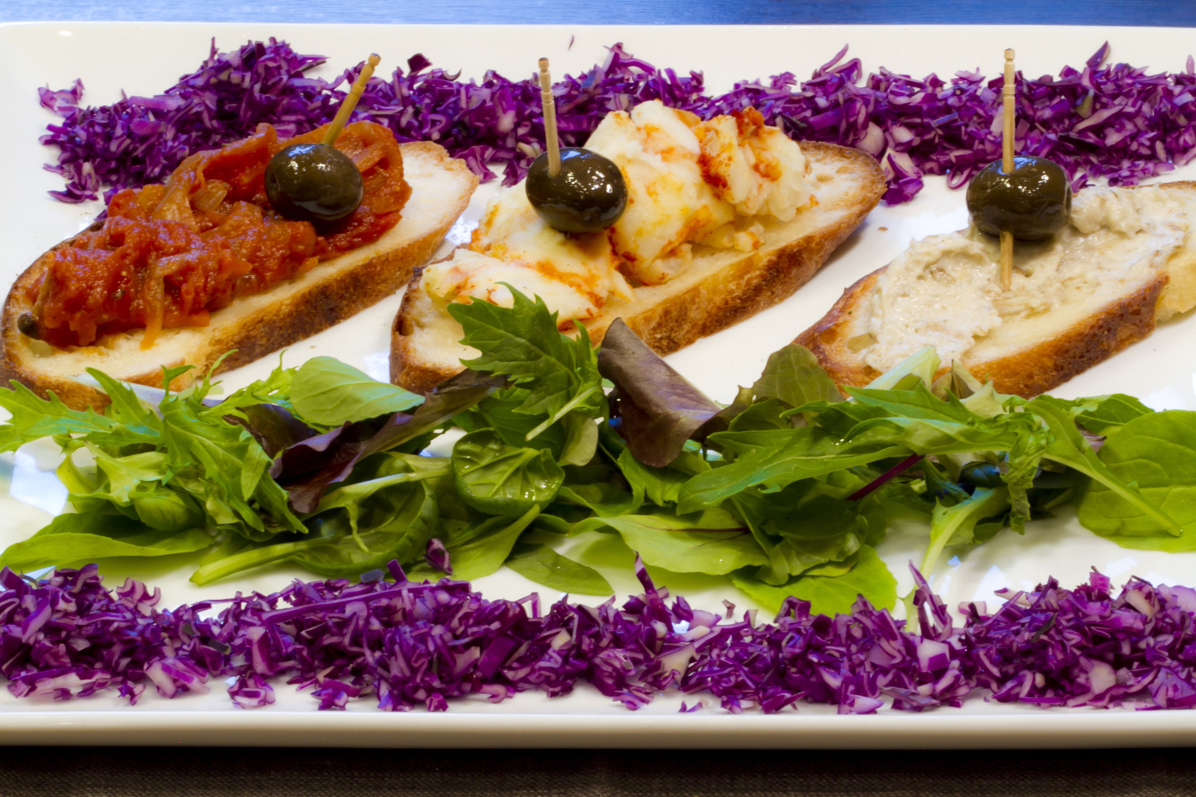 A plate with three pieces of bread topped with different spreads and olives, accompanied by a side of mixed greens and purple cabbage.