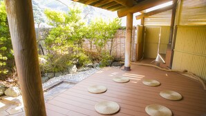 Una terraza de madera con esteras tejidas, rodeada por una valla de bambú y un sendero de piedra.