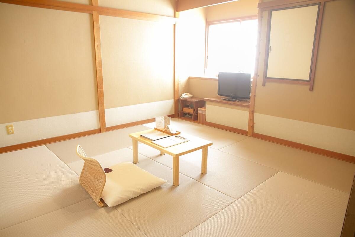 Una habitación japonesa tradicional con suelo de tatami, una mesa de madera baja, un cojín y un televisor al fondo.