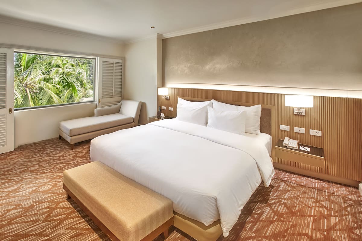 A hotel room with a large bed, a bench, a bedside table with a lamp, and a view of palm trees through the window.