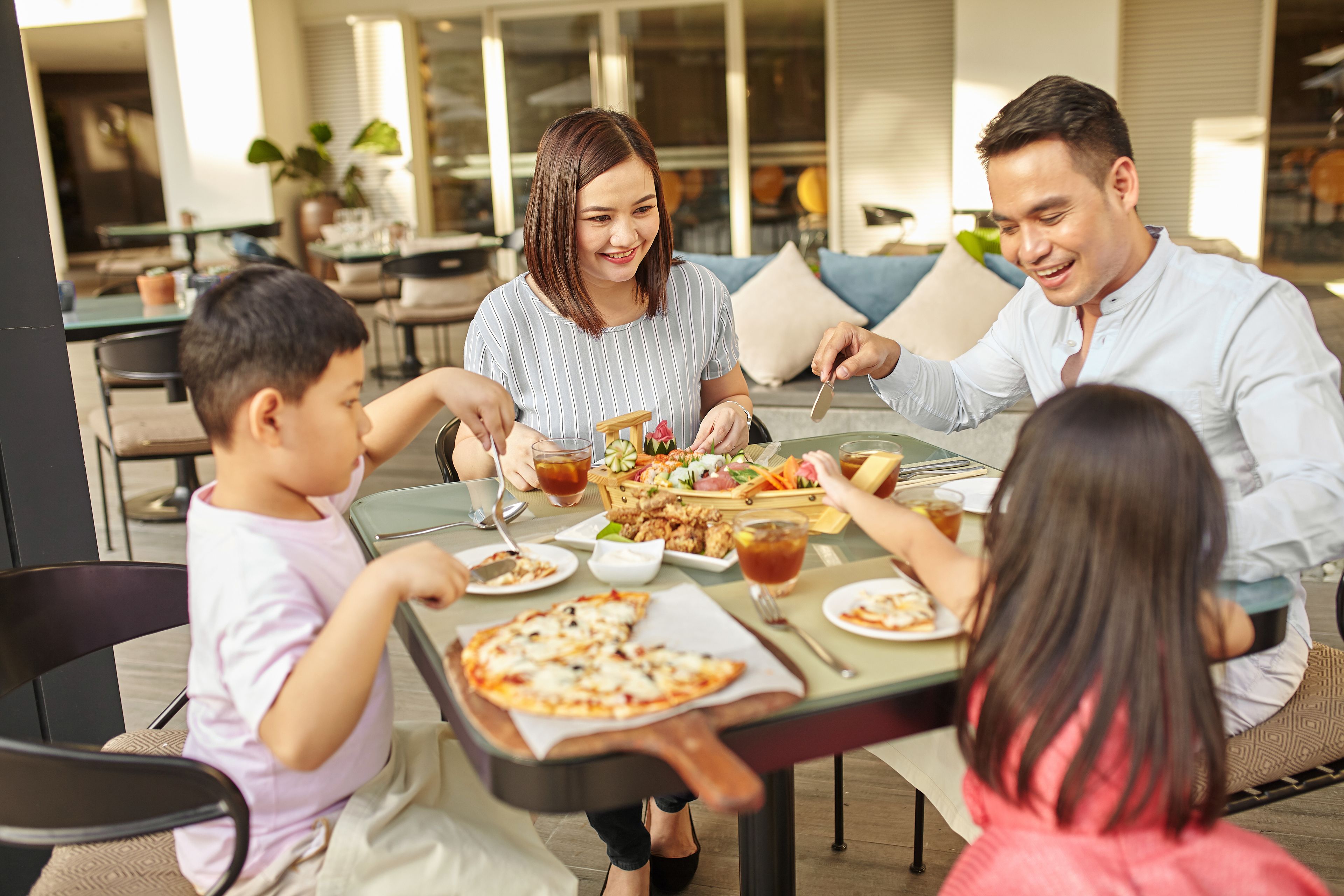 Una familia cena al aire libre en un restaurante, con diversos platos y bebidas sobre la mesa.
