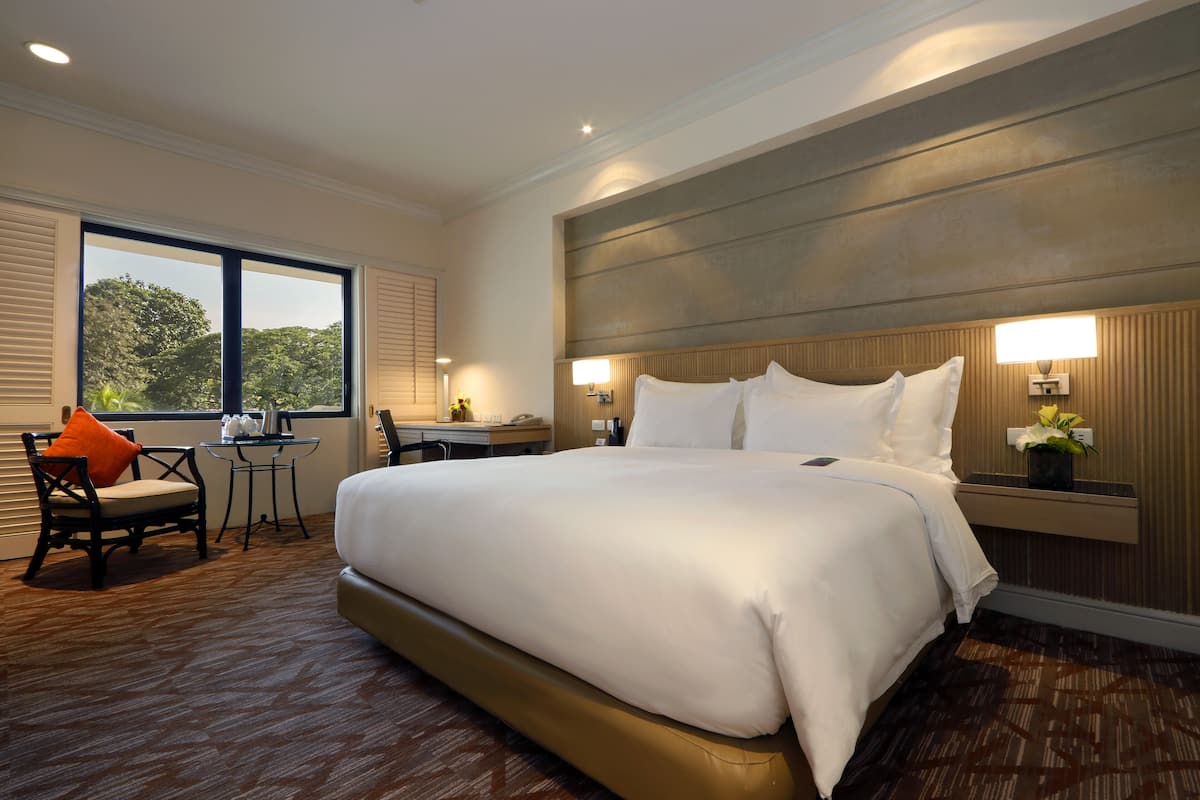 A large bed with white linens, a wooden headboard, bedside lamps, a small table with a chair, and a window with blinds.