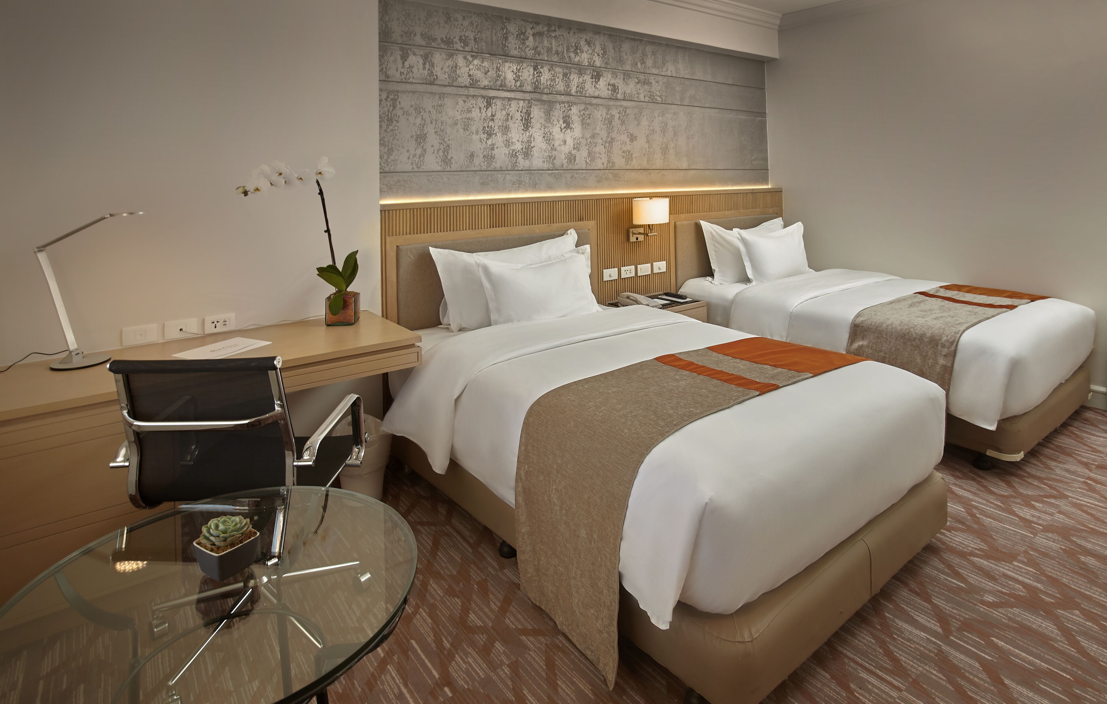 Ett hotellrum med två sängar, ett skrivbord med en lampa, ett glasbord med en växt och en dekorativ väggpanel.
