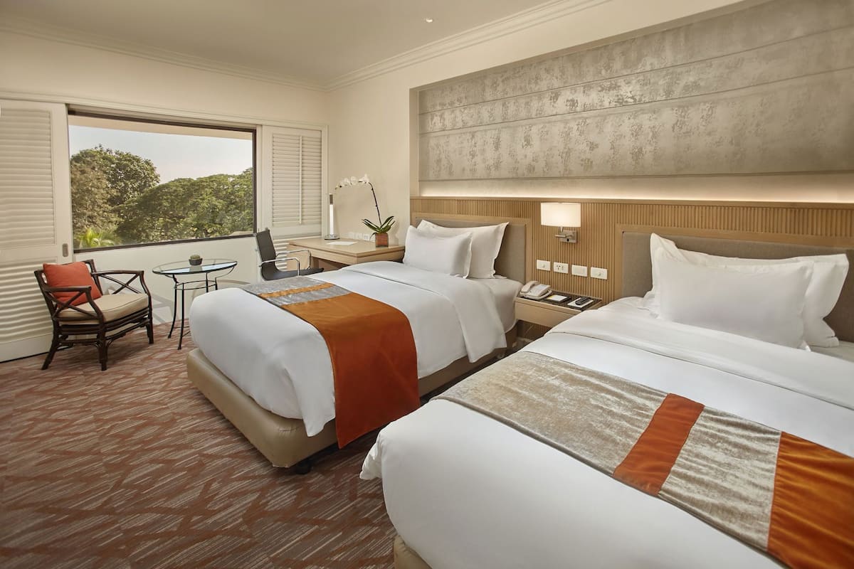 A hotel room with two beds, a desk, a chair, a small table, and a window with a view of trees.