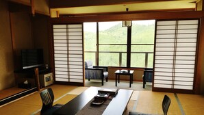 Una habitación japonesa tradicional con suelo de tatami, una mesa baja con cojines, un televisor y vistas a un paisaje verde exuberante a través de puertas shoji.