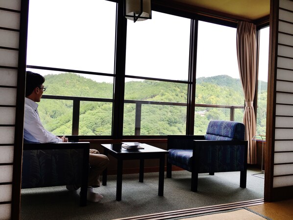 Habitación con un ventanal grande que ofrece vistas a un paisaje montañoso cubierto de bosque. Se observa una mesa y dos sillas, una de ellas con un cojín estampado.
