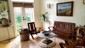 Un salon chaleureux avec un canapé en bois, une table basse ronde et une bibliothèque. On aperçoit une plante en pot sur une petite table et un tableau accroché au mur.