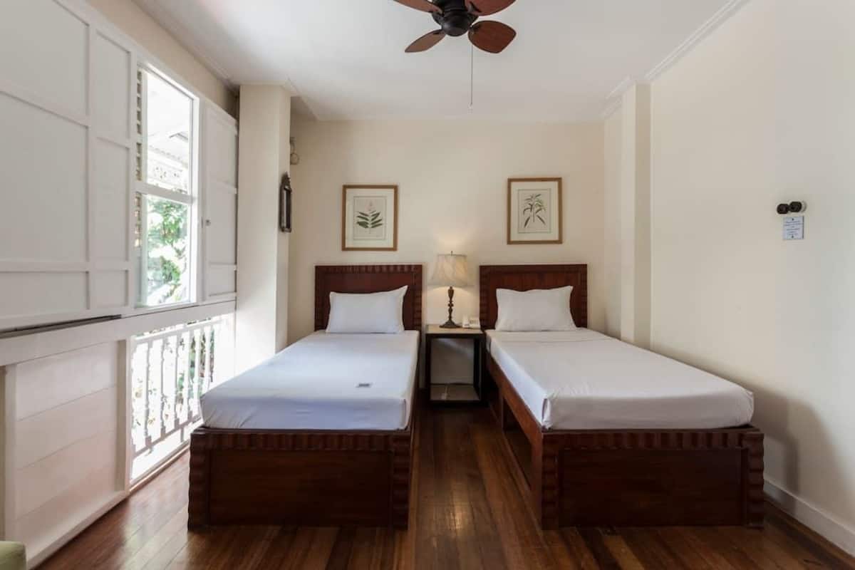 Une chambre avec deux lits en bois, un ventilateur de plafond, des reproductions encadrées de motifs botaniques accrochées au mur et une fenêtre donnant sur un paysage verdoyant.