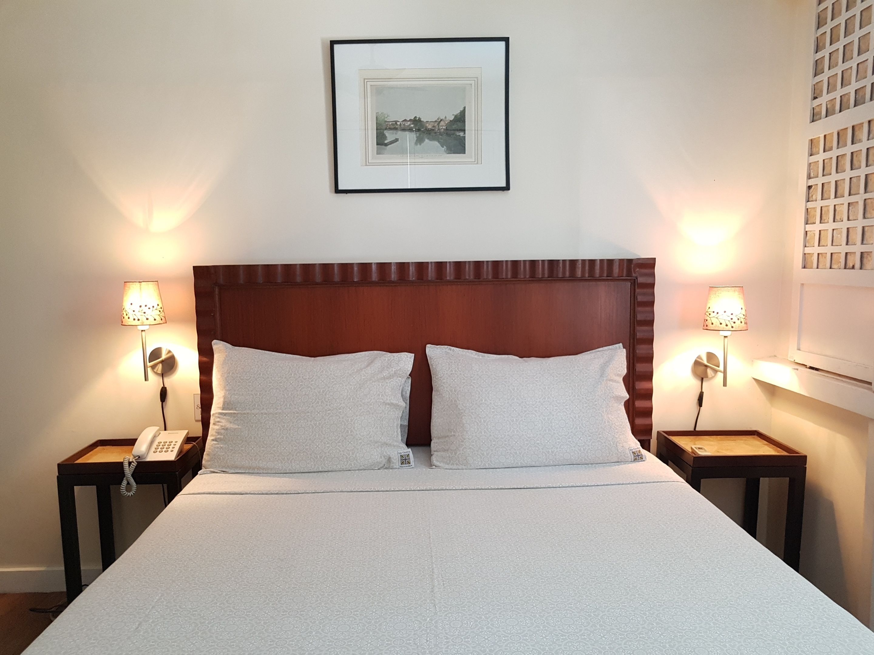 Een net opgemaakt bed met een houten hoofdbord, twee nachtkastjes met lampen, en een lijst met een afbeelding aan de muur.