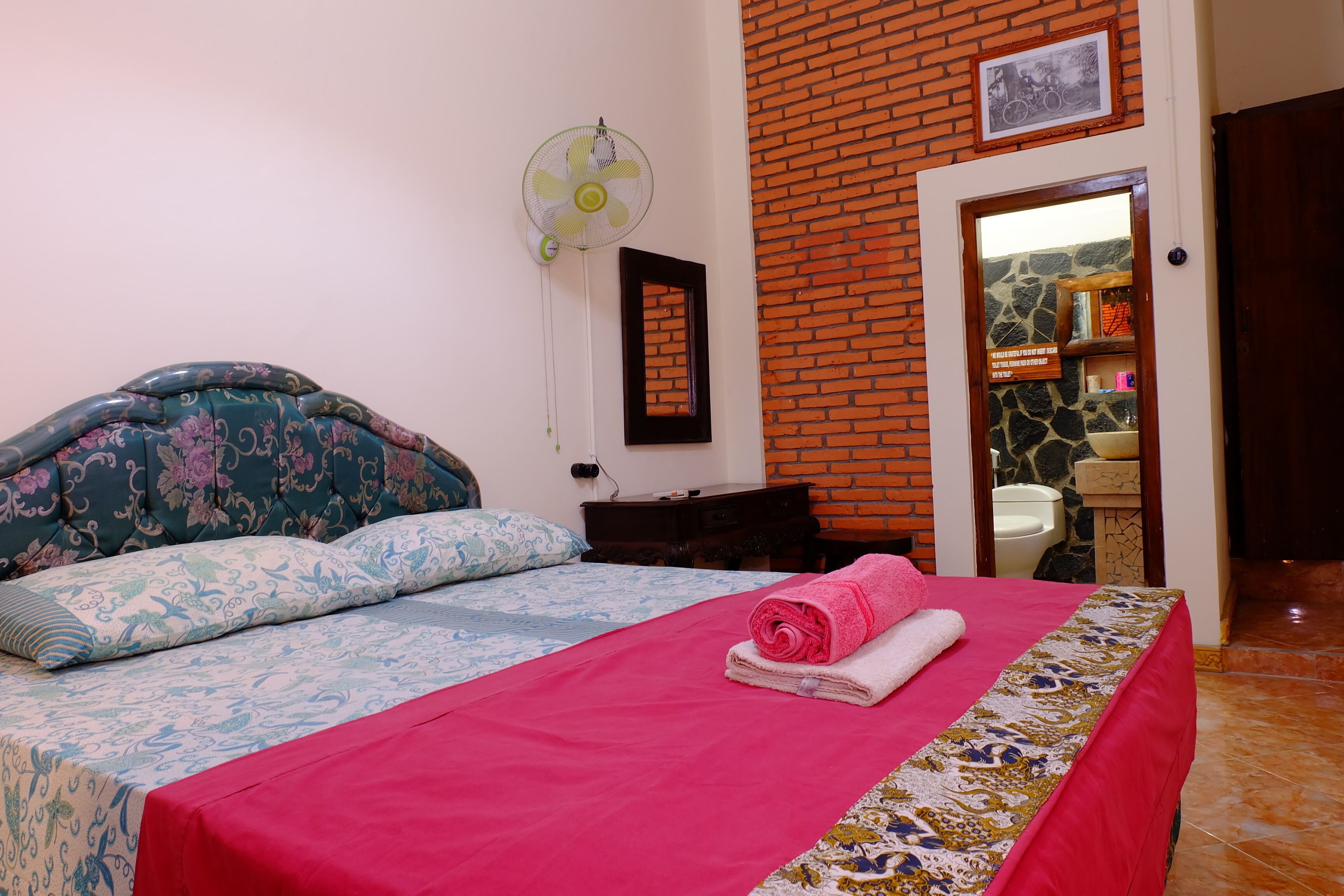 A hotel room with a bed, a fan, a mirror, and a bathroom visible through an open door.