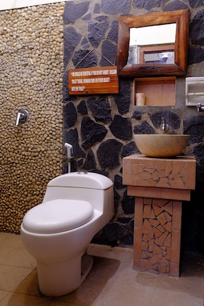 Un baño con pared de piedra, inodoro blanco, lavabo con cuenco y espejo con marco de madera.