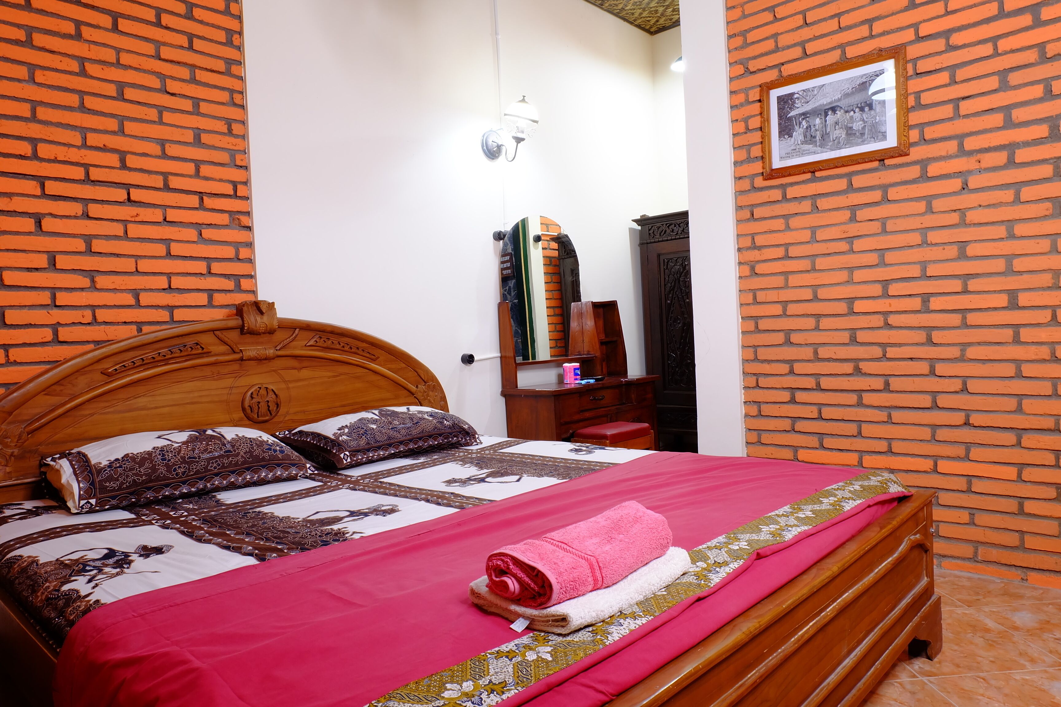 A neatly made bed with a red blanket and a pink towel, a wooden headboard, and a brick-patterned wall.