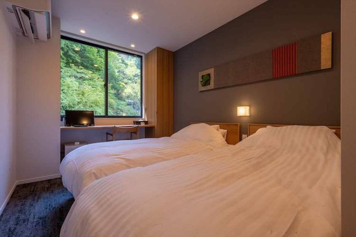A hotel room with a large bed, a desk with a computer, a window with a view of greenery, and a wall-mounted light.