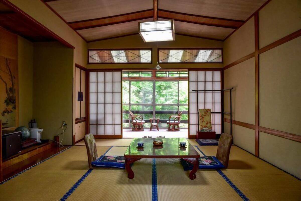 A traditional Japanese room with tatami flooring, a low table with cushions, sliding doors, and a view of greenery outside.