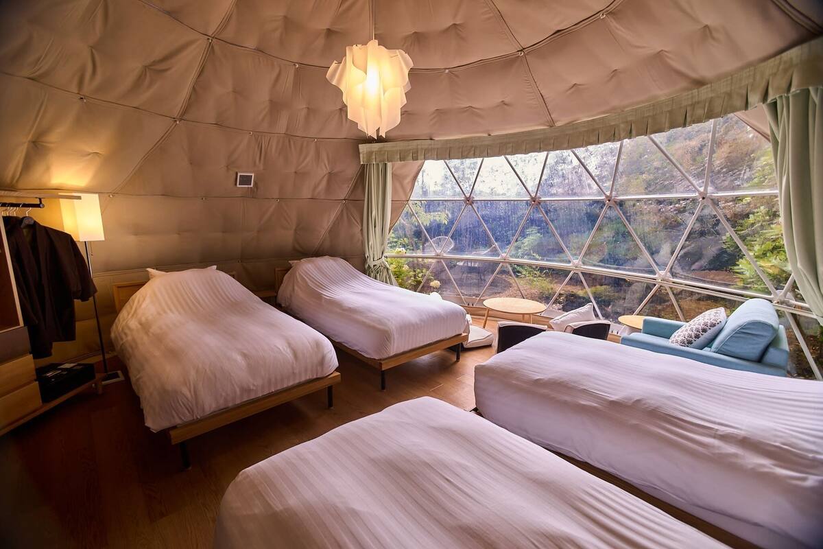 A tent-style room with two beds, a small table, and a view of a forest through a large window.
