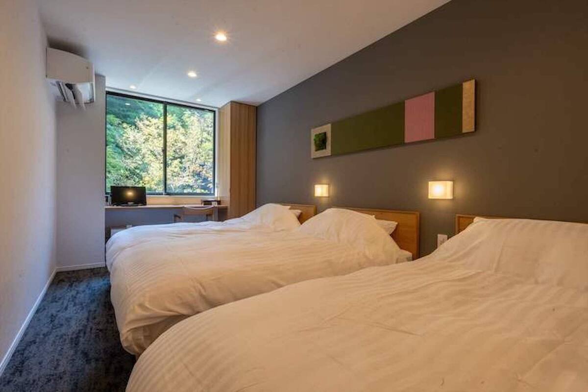 A hotel room with two beds, a desk, a window with a view of trees, and wall-mounted lights.