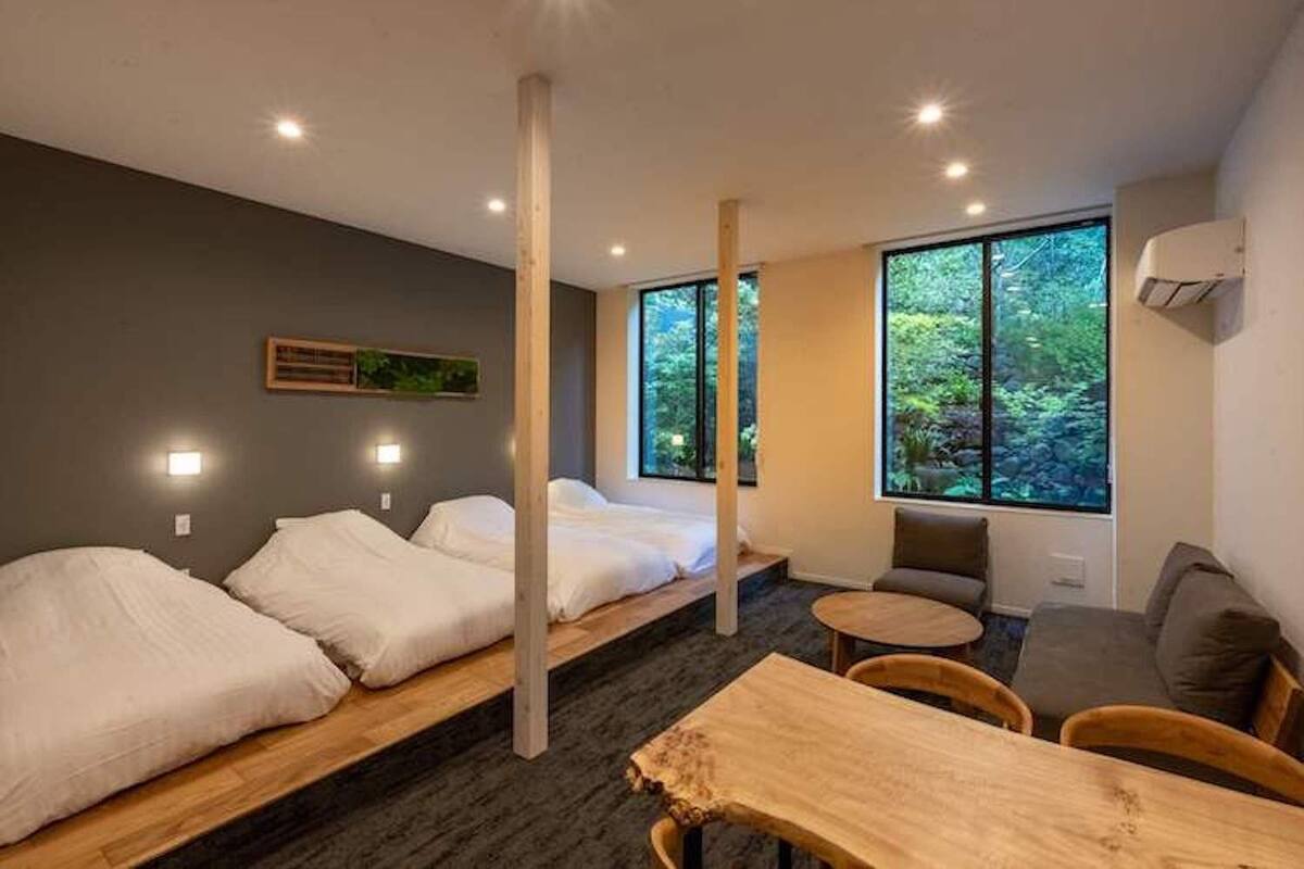 A modern hotel room with a bed, a small table, a chair, and large windows offering a view of greenery.