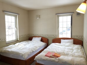 Habitación con dos camas, cada una con cabecero de madera y ropa de cama blanca. Hay ventanas a ambos lados de la habitación, y se ve una lámpara a la derecha.