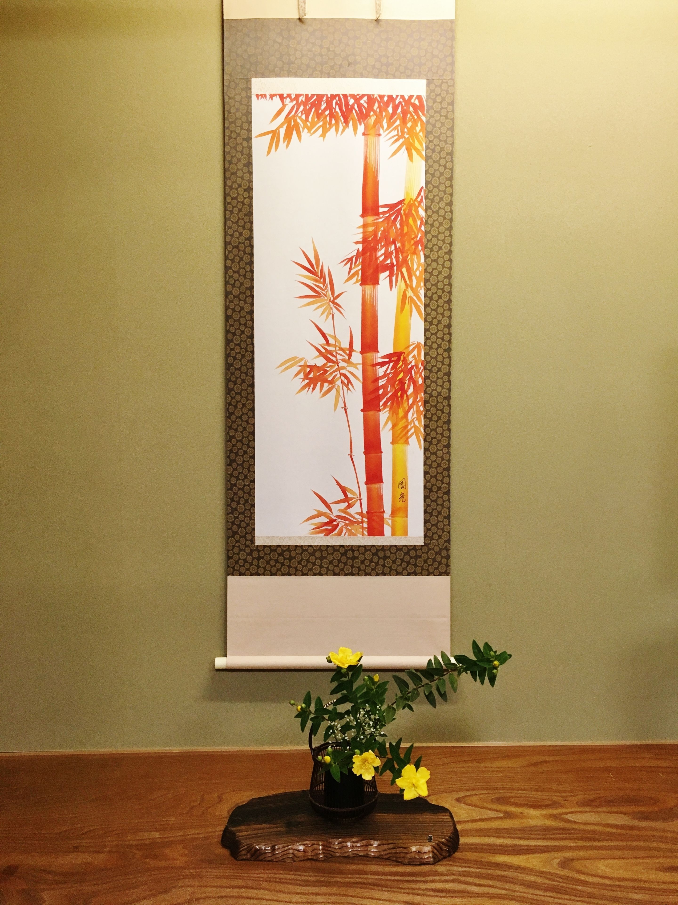 Une œuvre d’art encadrée représentant des bambous, accrochée à un mur, une table en bois sur laquelle est posé un vase rempli de fleurs jaunes, et un mur orné de motifs.