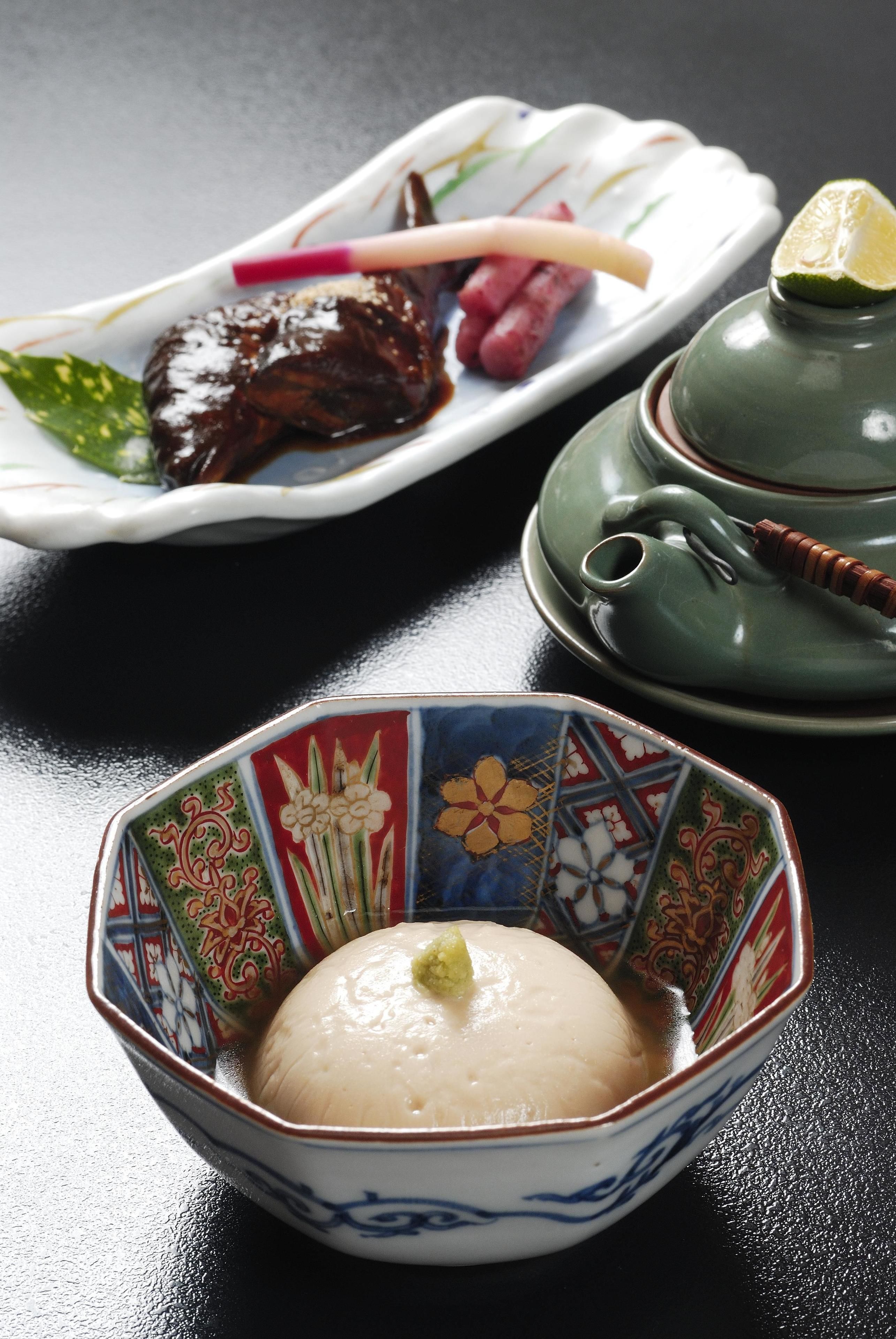 A bowl of white rice with a green garnish, a plate with a piece of meat and vegetables, and a green teapot on a black table.