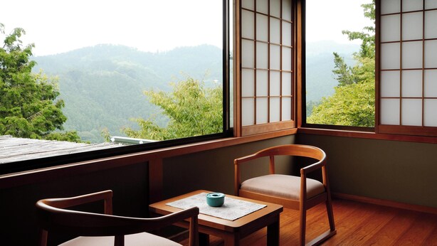 Una habitación japonesa tradicional con suelo de tatami, una mesa baja con un cuenco azul, dos sillas de madera y vistas a un paisaje montañoso a través de paneles shoji.