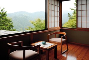 A traditional Japanese room with tatami flooring, a low table with a blue bowl, two wooden chairs, and a view of a mountainous landscape through shoji screens.