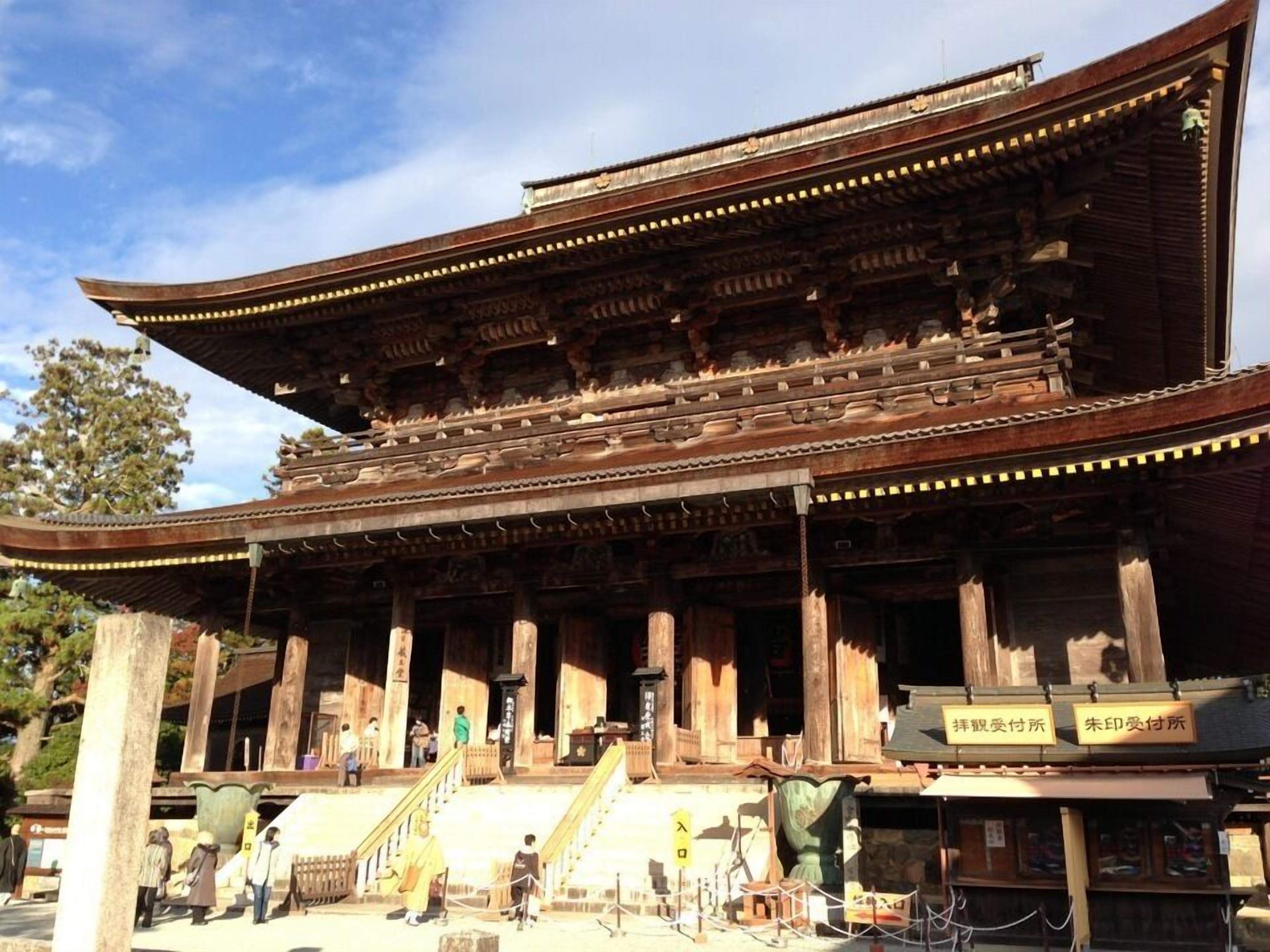 A traditional wooden building with a curved roof and multiple columns. There are signs in a foreign language, and people are seen walking around.