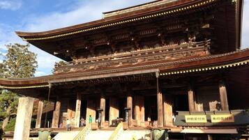 A traditional wooden building with a curved roof and multiple columns. There are signs in a foreign language, and people are seen walking around.