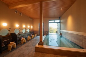 A modern spa with a large indoor pool, wooden benches, and multiple mirrors.