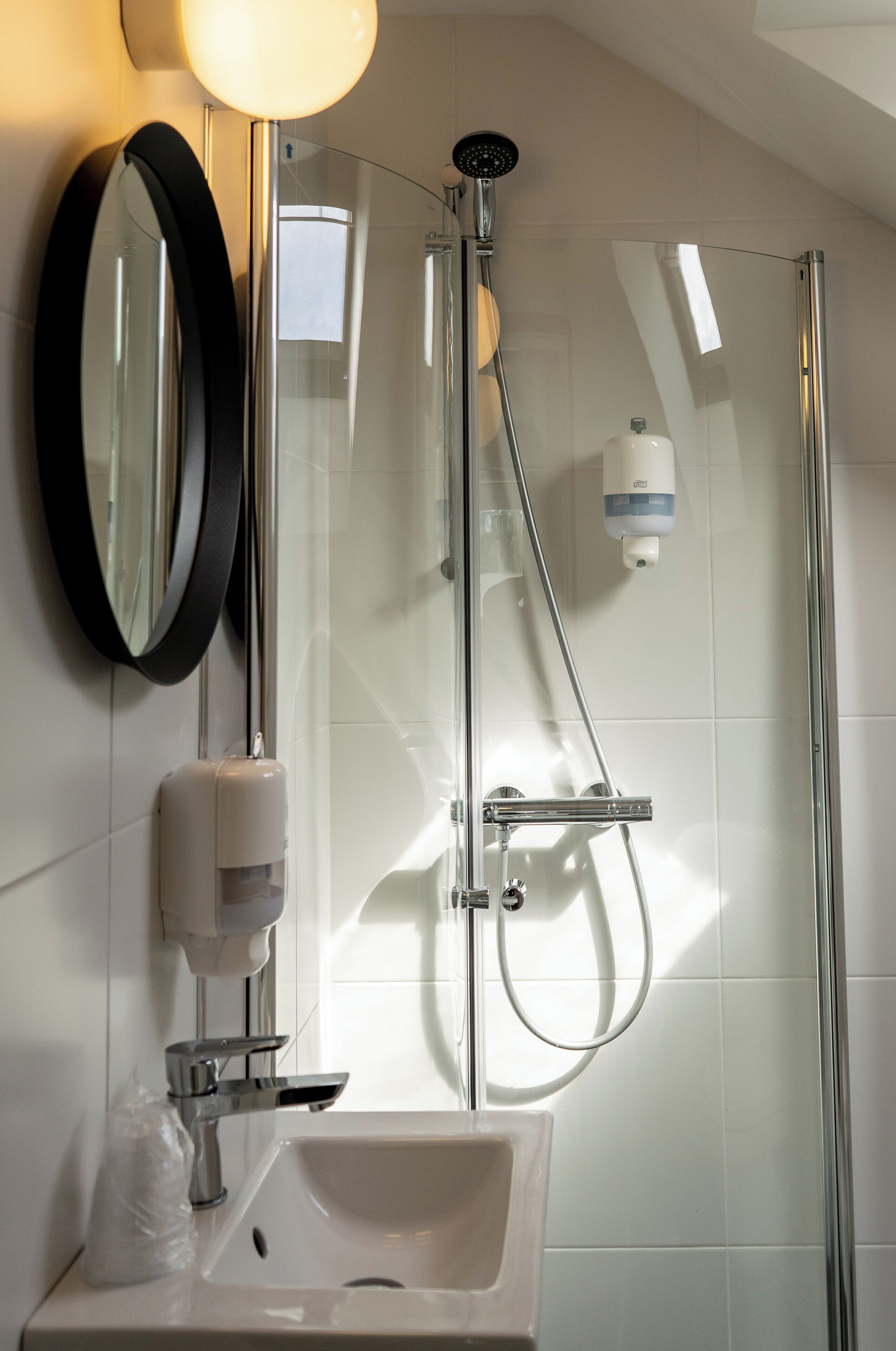 Un baño moderno con ducha envuelta en vidrio, un lavamanos blanco, un espejo y un dispensador de jabón.