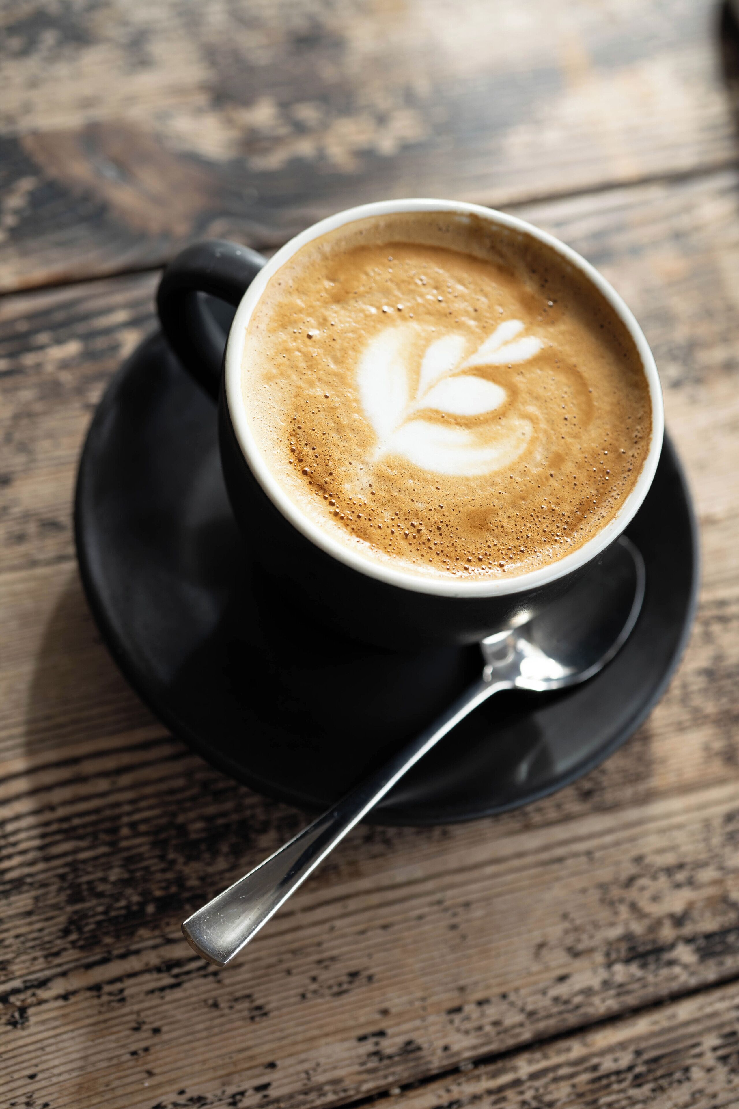 A cup of coffee with latte art on a saucer and spoon, placed on a wooden table.