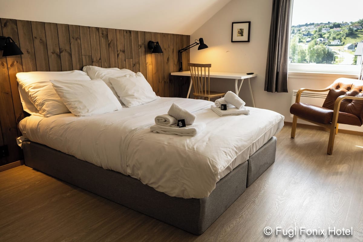 Una cama bien hecha con ropa blanca, una mesita con una silla, y vista al exterior a través de una ventana.