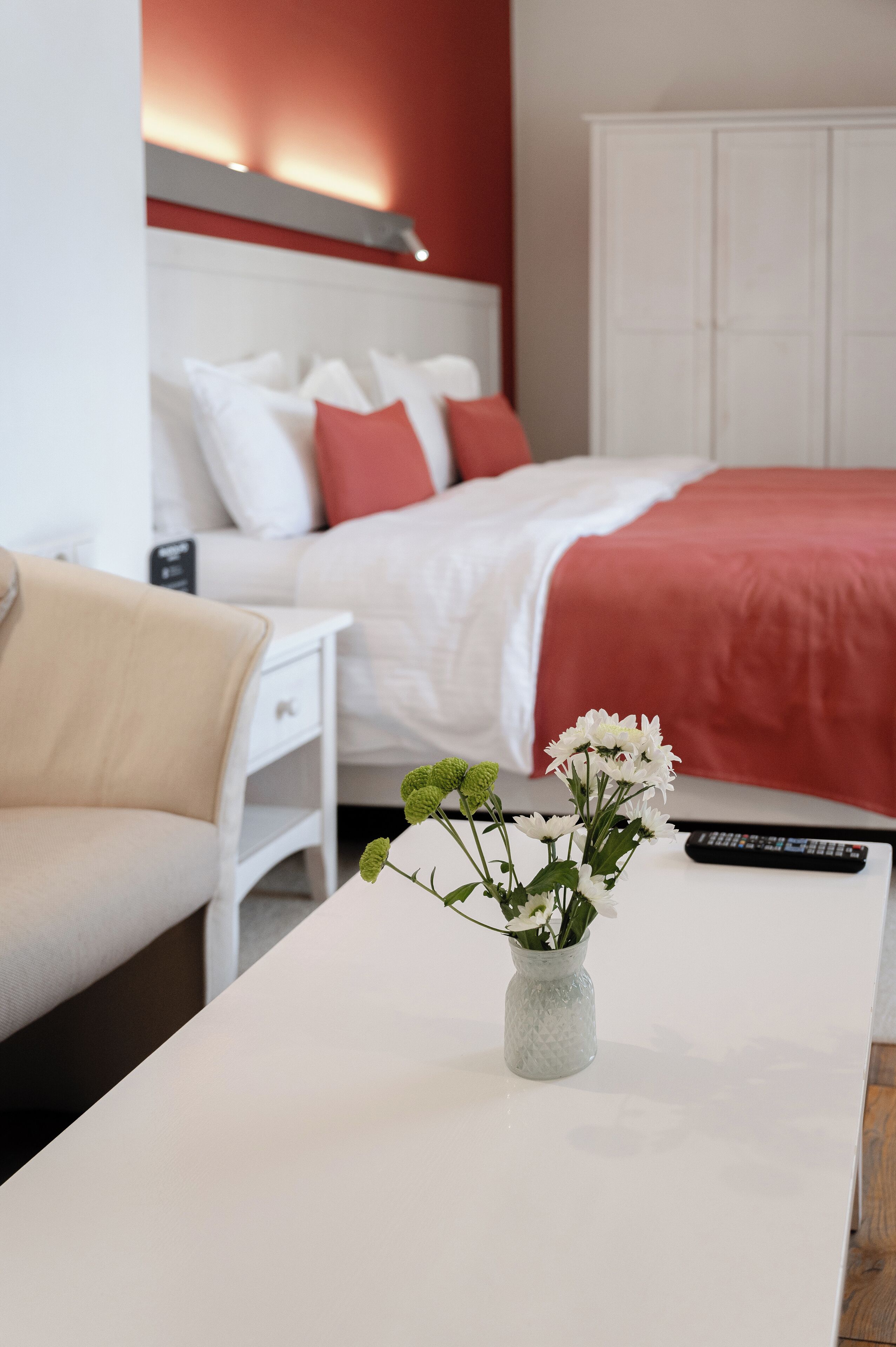 A neatly arranged hotel room with a bed, a white nightstand, a beige armchair, and a white table with a vase of flowers.