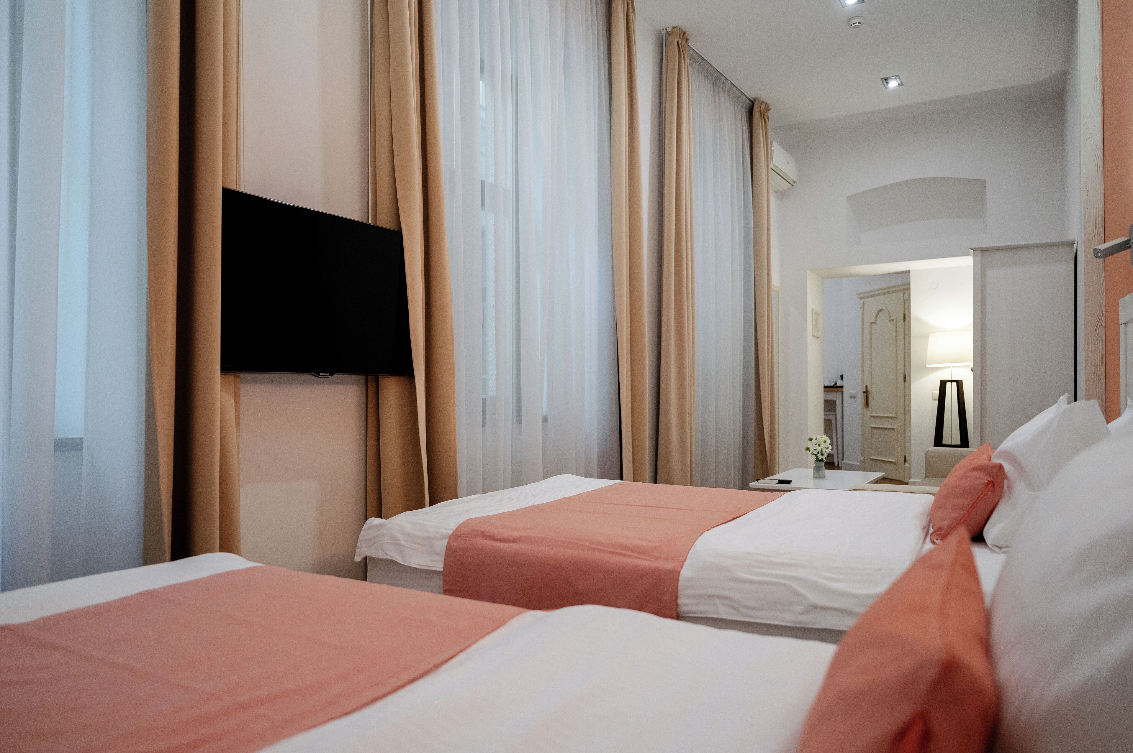 A hotel room with two beds, beige curtains, a flat-screen TV mounted on the wall, and a door leading to a bathroom.