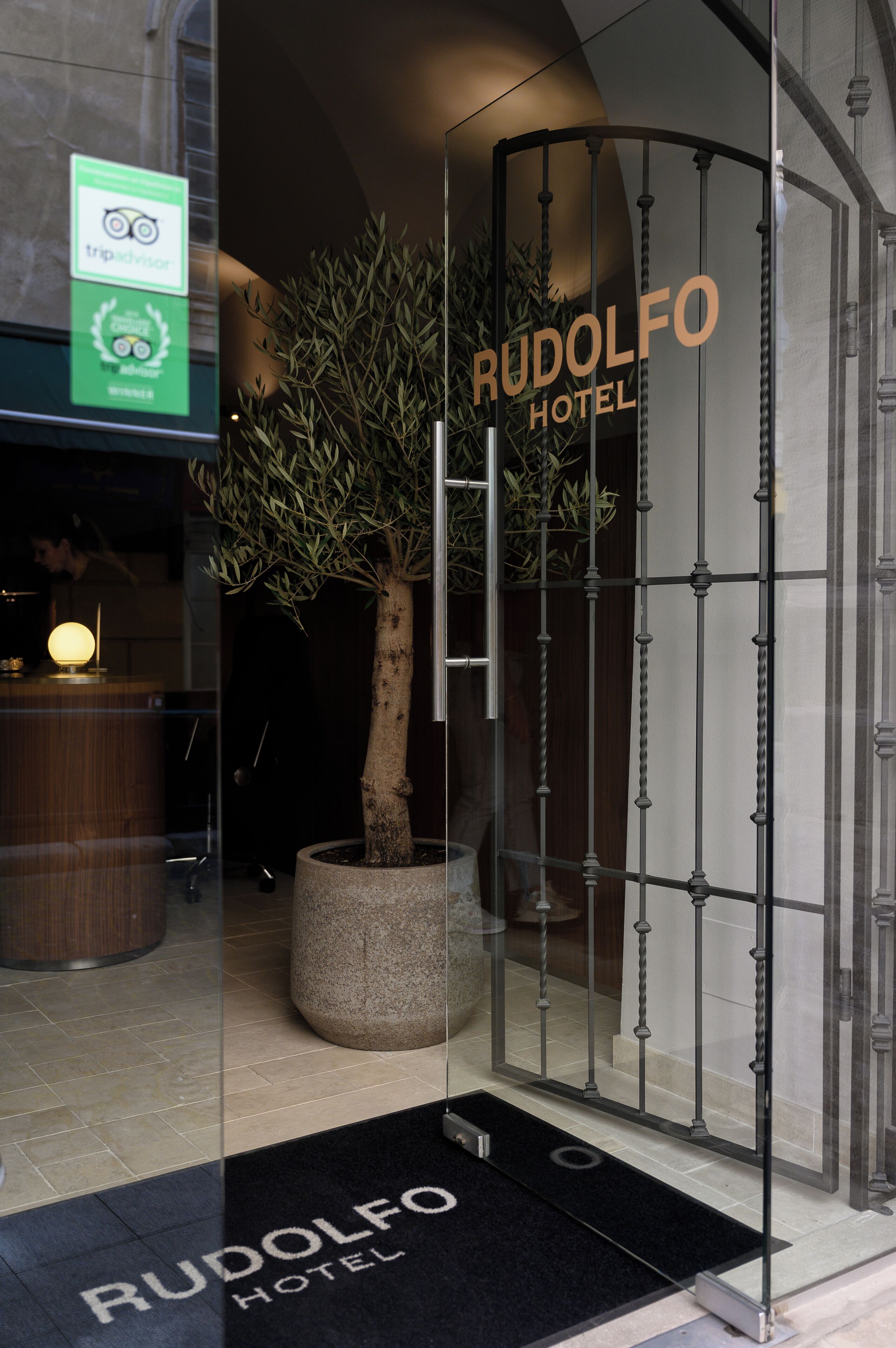 A glass entrance to Rudolf Hotel with a potted tree and a reception desk.