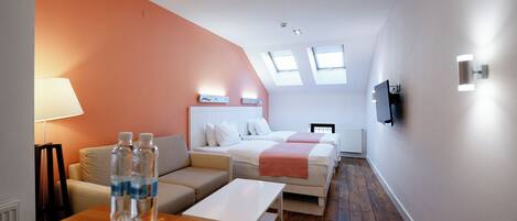 A hotel room with a sofa, two beds, a TV, and a table with water bottles.
