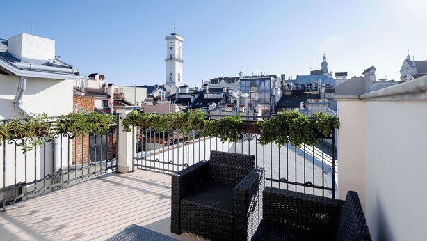 Una terraza en la azotea con mobiliario de mimbre, una valla metálica y vistas de un paisaje urbano que incluye una torre con reloj.