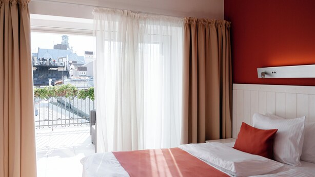 A hotel room with a large bed, a red throw blanket, white pillows, and a view of buildings through sheer curtains. - Rudolfo  Hotel (Lviv)