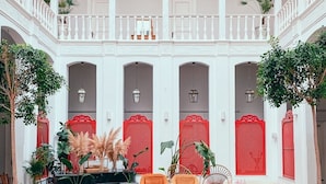 Un amplio patio con una larga mesa de madera rodeada de sillas de mimbre, paneles decorativos rojos y plantas en macetas.