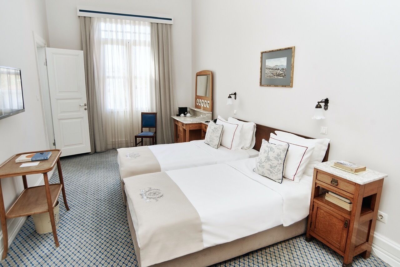 A hotel room with two beds, a desk, a chair, a small table, a nightstand, a mirror, a picture on the wall, and a window with curtains.