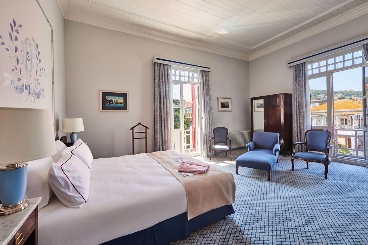 A hotel room with a bed, two armchairs, a desk with a lamp, and a view of the city through large windows.