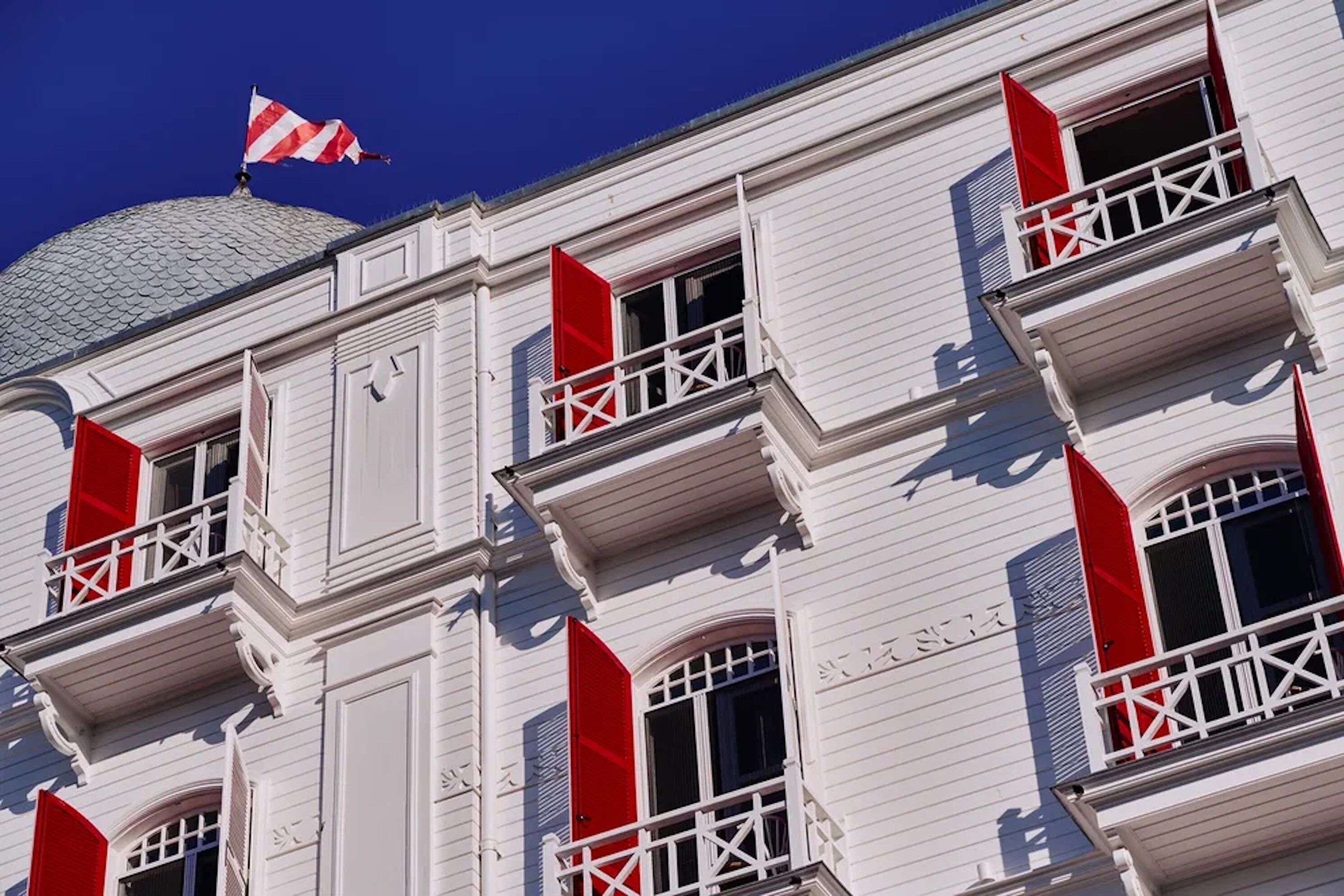 Un immeuble aux murs extérieurs blancs, doté de nombreux balcons ornés d’auvents rouges et surmonté d’un drapeau.
