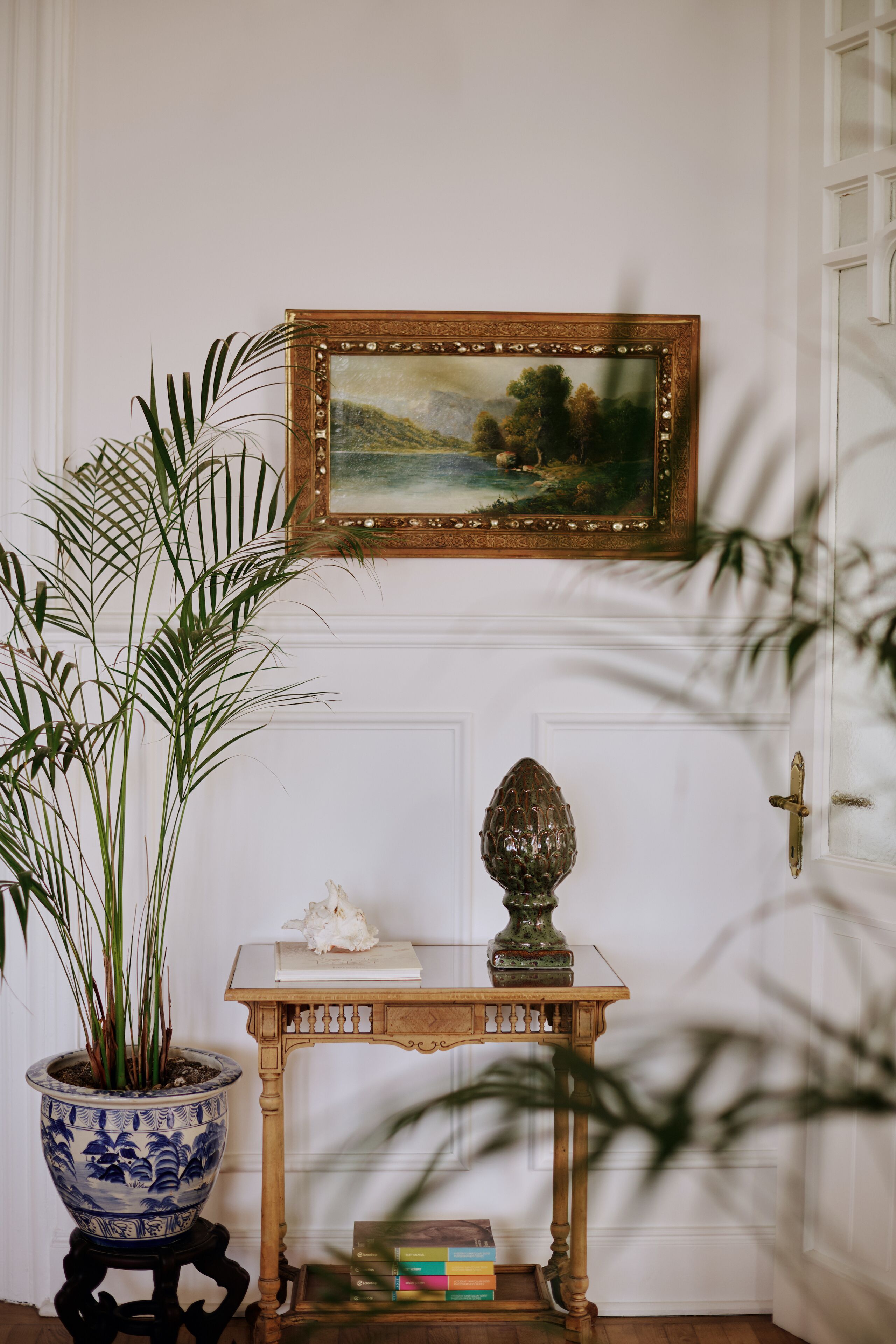 A room with a painting, a potted plant, a decorative vase, and a small table with books.